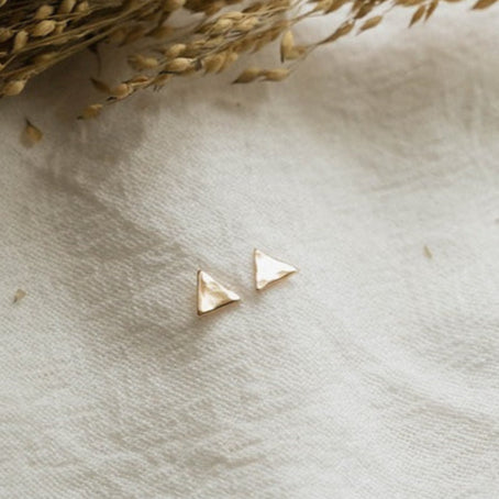 Gold triangle earrings on a textured beige surface with dried grasses.