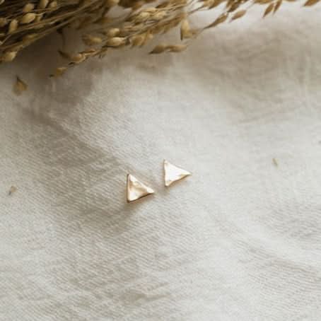 Gold triangle earrings on a textured beige surface with dried grasses.