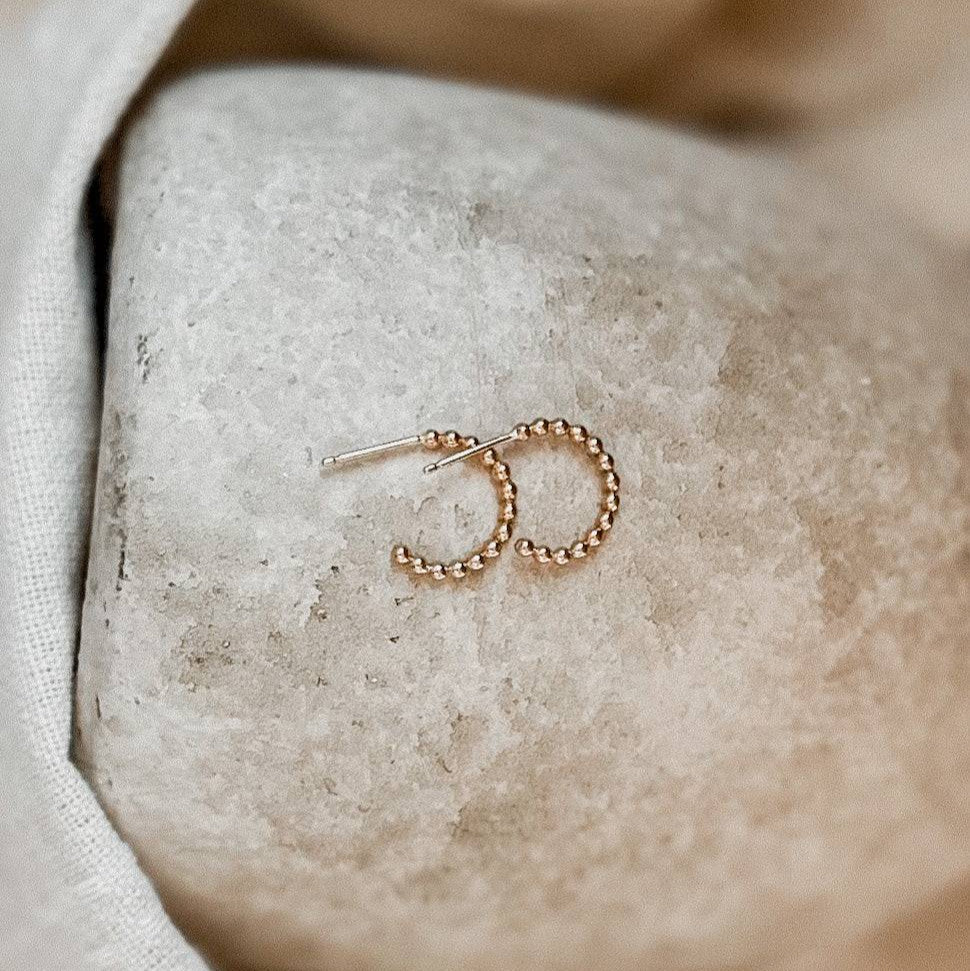 Beaded Huggie Hoop Earrings placed on a stone surface. Delicate 14k gold fill hoops designed for sensitive skin and daily wear.