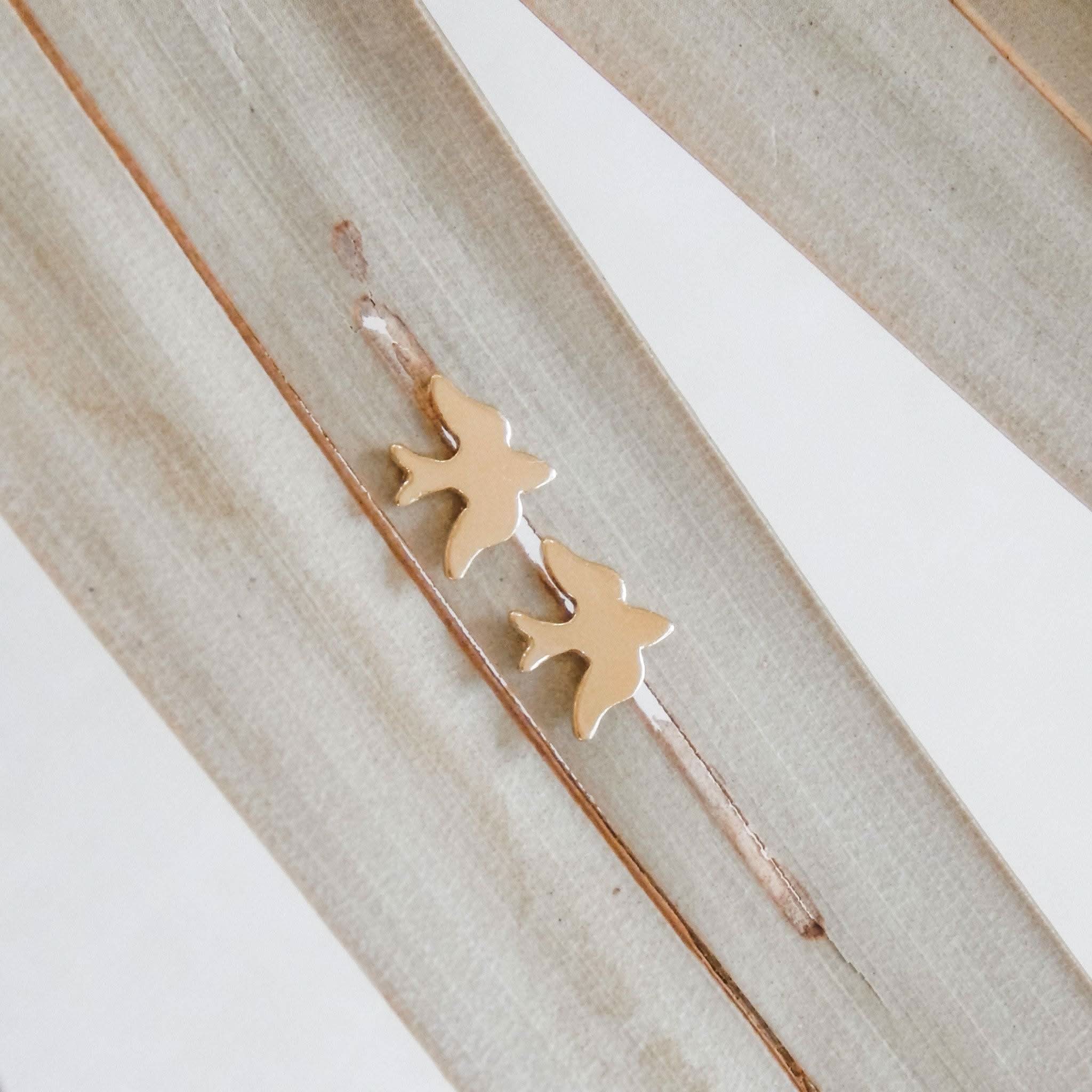 Petite Bird Stud Earrings in 14k Gold Fill from Barberry & Lace Jewelry, displayed on neutral-toned palm leaves for a minimal aesthetic.