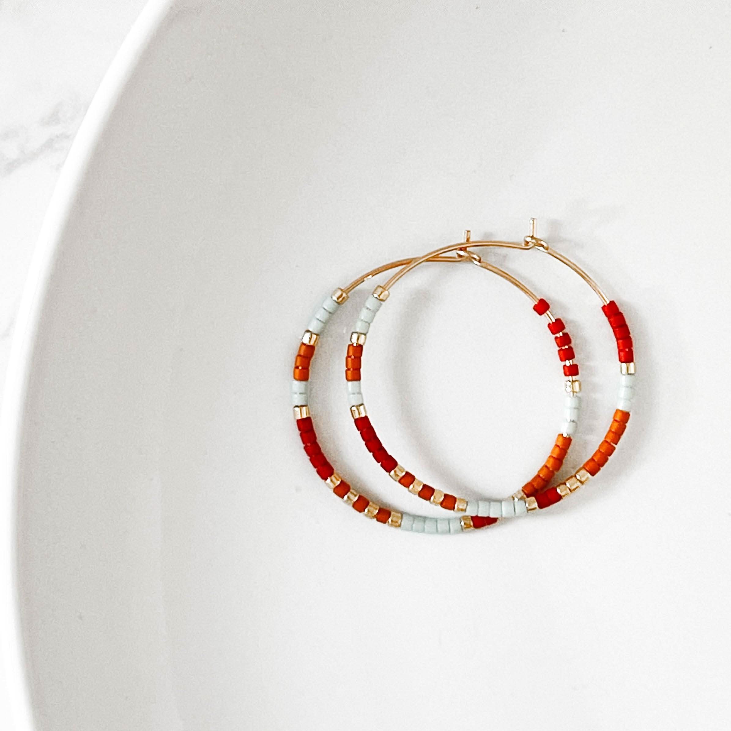 Desert Sunrise colorway of Summer Edition - Beaded Hoop Earrings with red, orange, and light blue seed beads on gold hoops, flat lay on white plate.