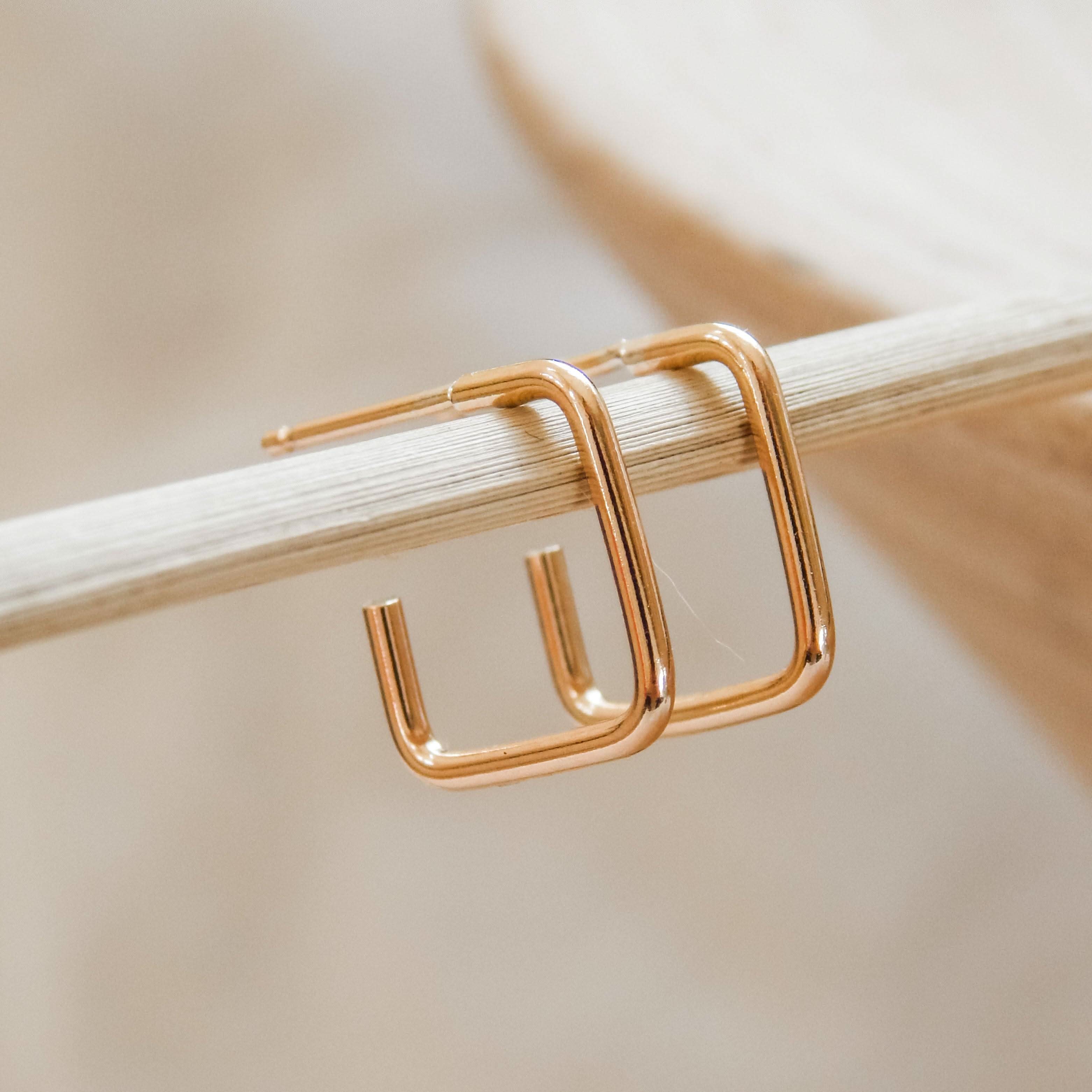 Square Hoop Stud Earrings in 14k gold fill with a sleek geometric design, displayed on a wooden rod. Modern and minimalist jewelry style.