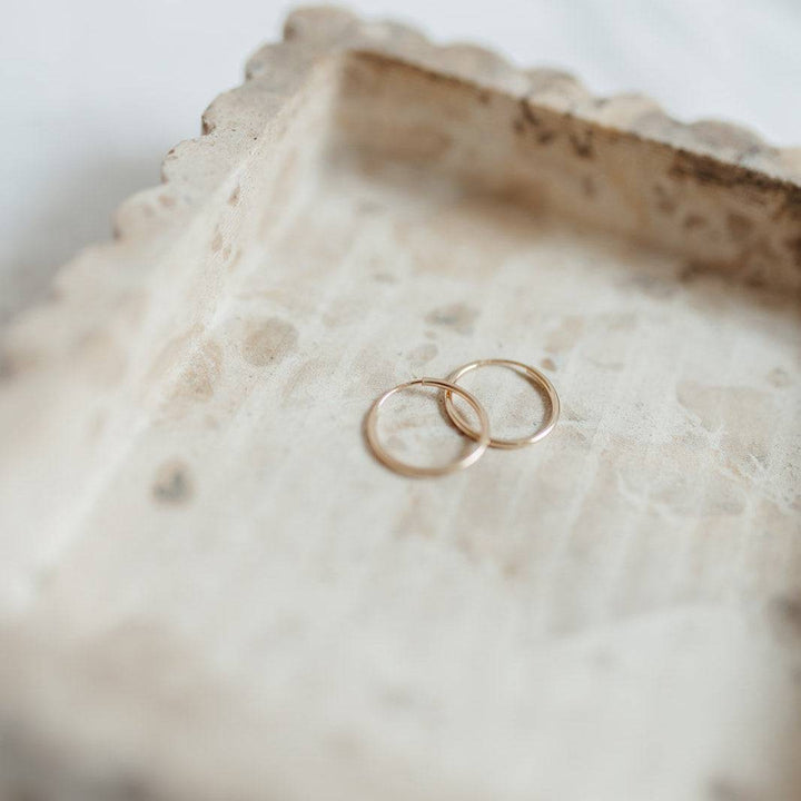 Pair of Endless Huggie Hoop Earrings in 14k gold fill displayed on a rustic stone tray. Minimalist and hypoallergenic jewelry for daily wear.