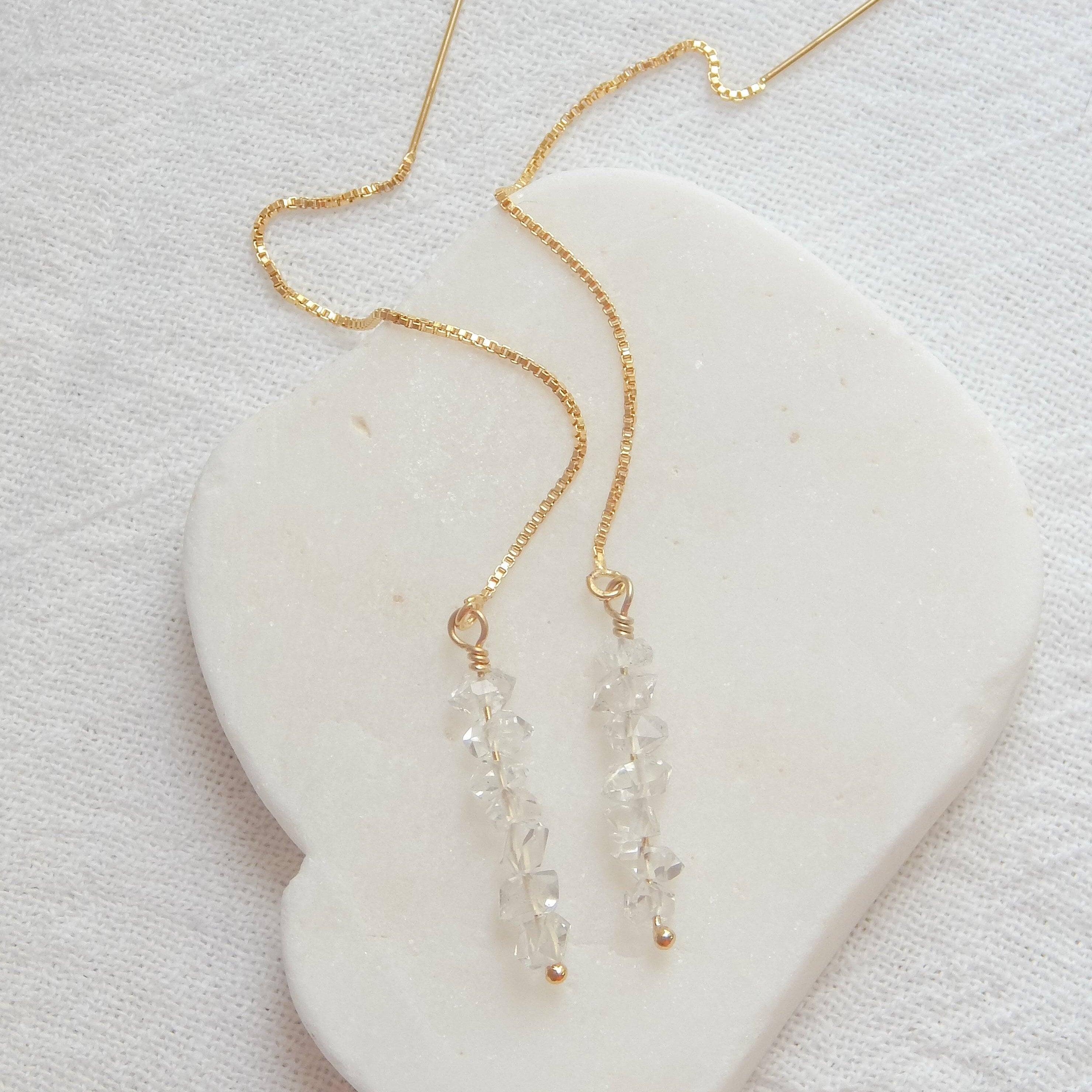 Flat lay of Herkimer Diamond Threaded Earrings on a white heart-shaped stone. Elegant and minimalistic gold and crystal design.