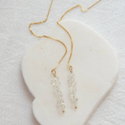 Flat lay of Herkimer Diamond Threaded Earrings on a white heart-shaped stone. Elegant and minimalistic gold and crystal design.