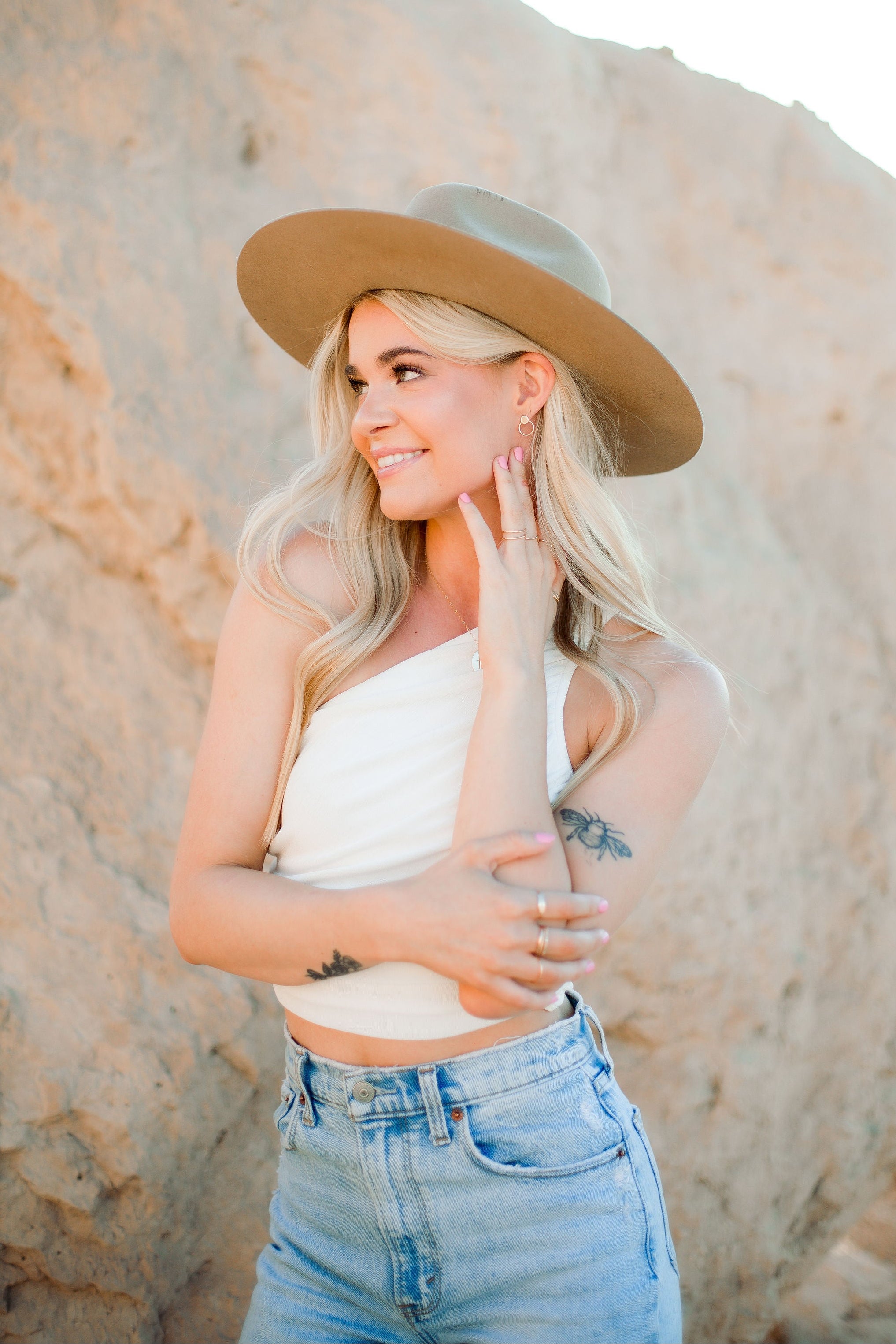 Woman wearing a beige hat, white top, and blue jeans standing against a rock formation.