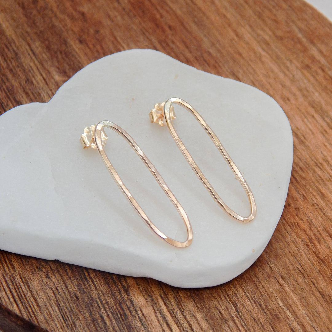 Hammered oval stud earrings in gold on a white stone and wooden backdrop.