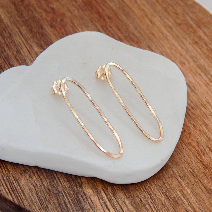 Hammered oval stud earrings in gold on a white stone and wooden backdrop.