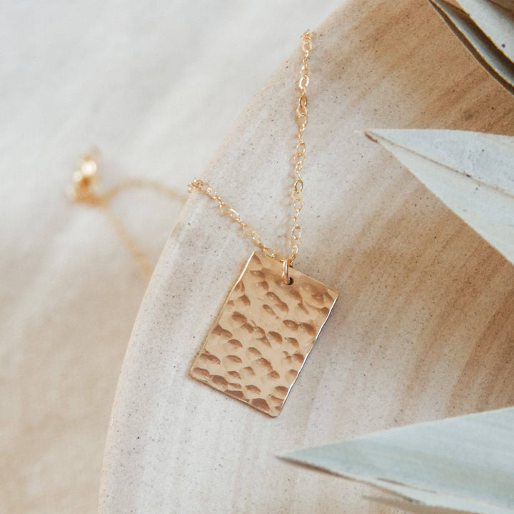 Hammered Rectangle Necklace by Barberry & Lace Jewelry displayed on a ceramic dish. Features a textured 14k gold fill pendant on a delicate chain.