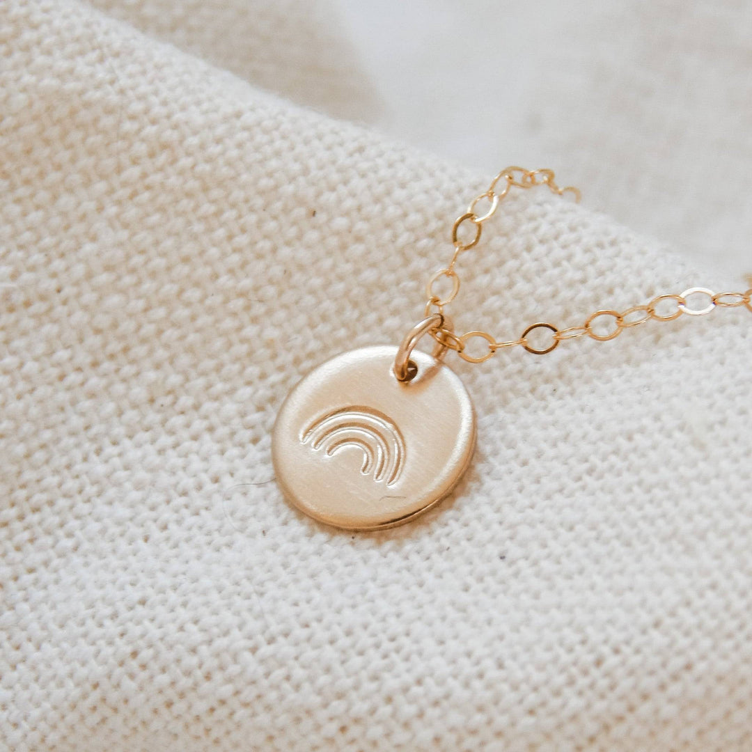 Hand stamped rainbow disc necklace in 14k gold fill with delicate chain, displayed on neutral fabric. Hypoallergenic and waterproof jewelry.