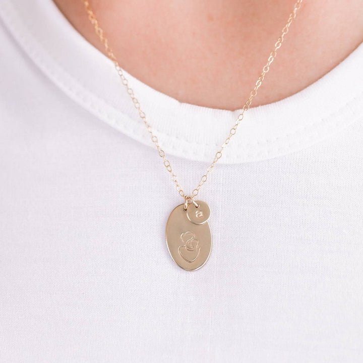 Hand Stamped Mother & Child Necklace by Barberry & Lace Jewelry featuring a gold oval pendant with engraved mother and child design and customizable initial charm.